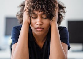 Woman in office setting expresses stress, seated at desk with hands in hair. Perfect for workplace stress concept.