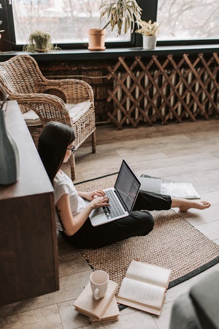 A young woman in casual attire working from home using a laptop while sitting on the floor with a coffee mug nearby.