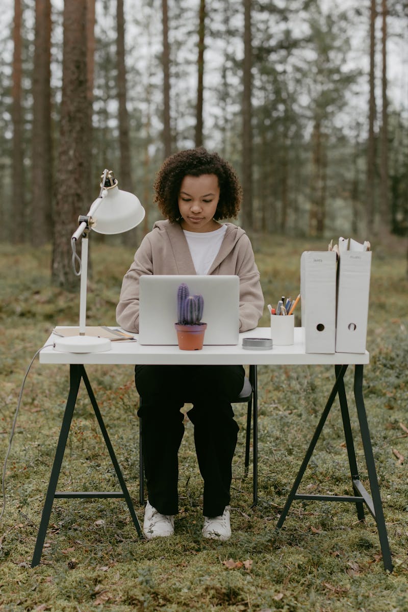 A woman sits at a desk in a forest, working remotely on a laptop amidst nature.